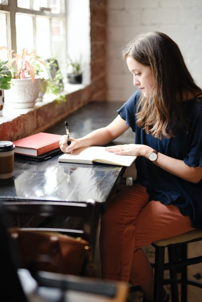 woman sitting in front of black table writing on white book near window